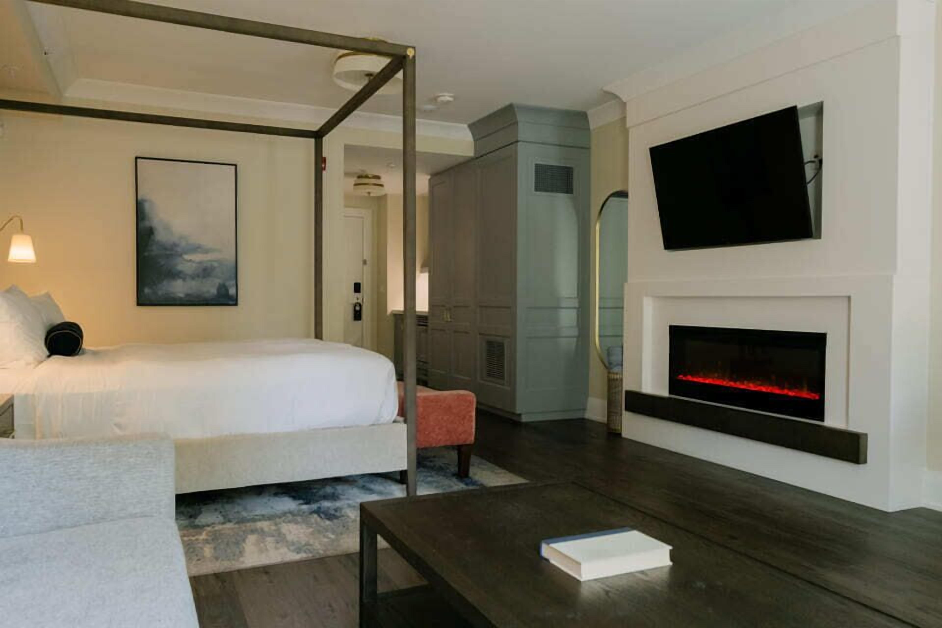 A modern hotel room with a four-poster bed, a flat-screen TV mounted above a fireplace, and a cozy seating area with a small table and chair.