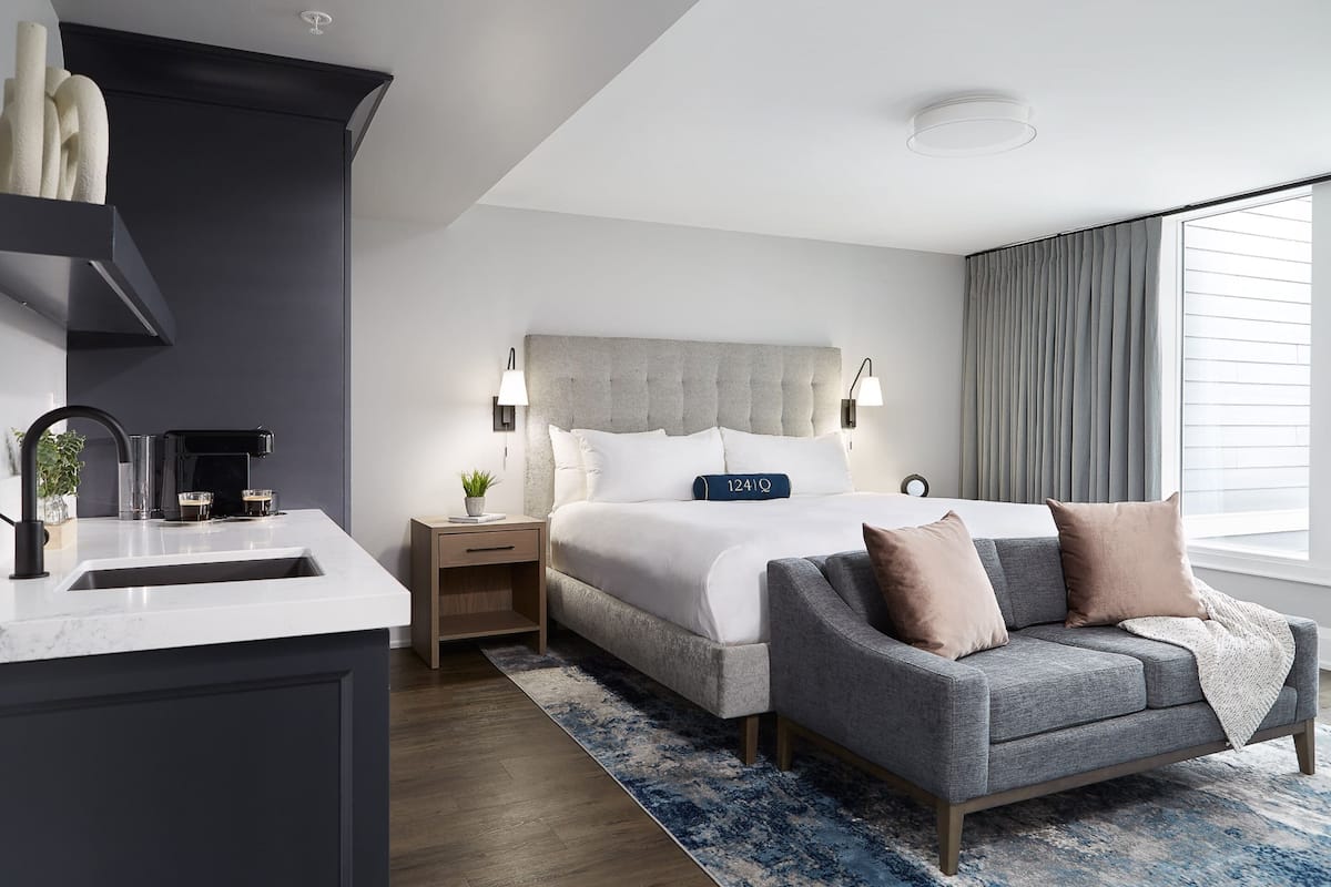 Une chambre d’hôtel moderne dotée d’un grand lit, de tables de chevet, d’un canapé, d’un comptoir en marbre et d’une grande fenêtre avec des rideaux.