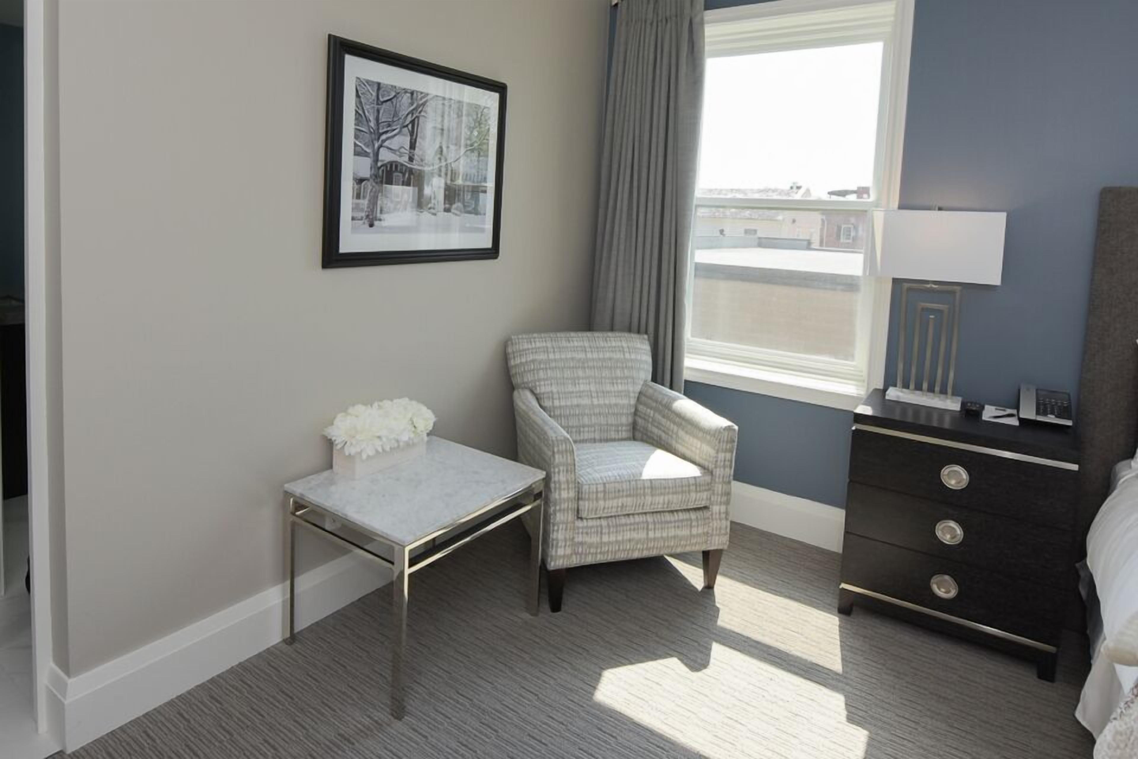Une chambre d’hôtel avec un lit, une chaise, une petite table, une commode, un tableau encadré au mur et une fenêtre avec des rideaux.