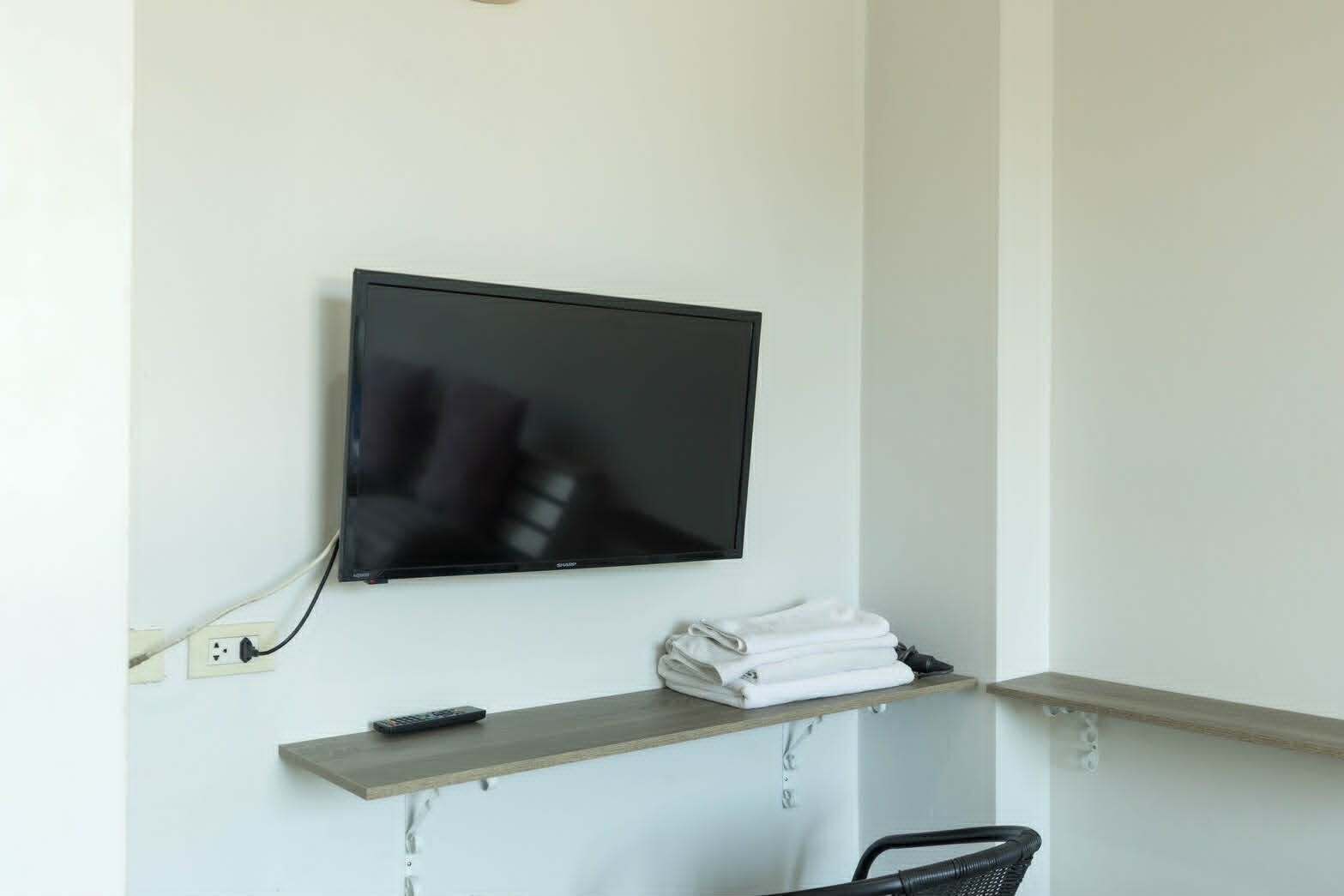 A TV mounted on a wall, a remote control, and a shelf with folded towels.