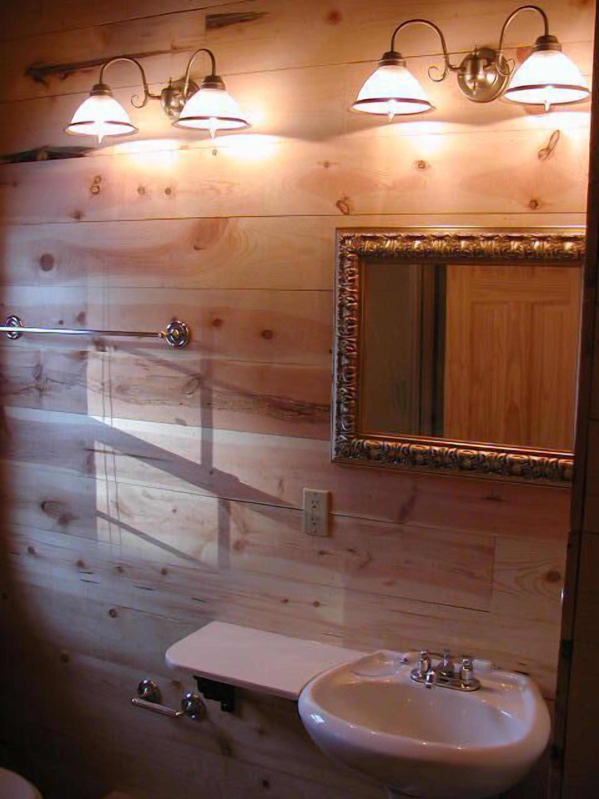 A wooden cabin bathroom with a white sink, a mirror with an ornate frame, and three wall-mounted lights.