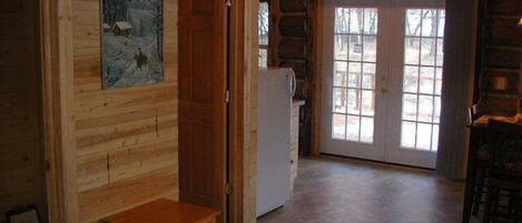 Interior de una cabaña de troncos con pared de madera, una puerta, un refrigerador, una ventana y una pequeña mesa de madera.