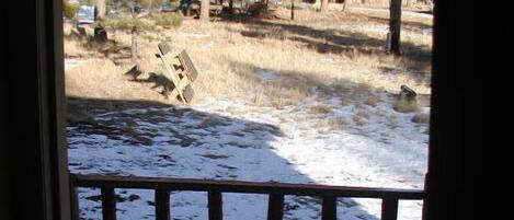 A view from a window with a wooden railing, overlooking a snowy landscape with trees and a fallen object.