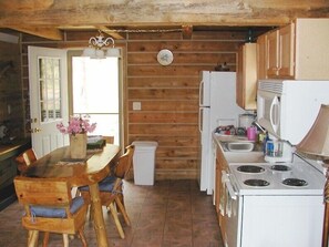 A rustic kitchen with wooden walls, a wooden dining table with chairs, a refrigerator, a stove, and a sink.
