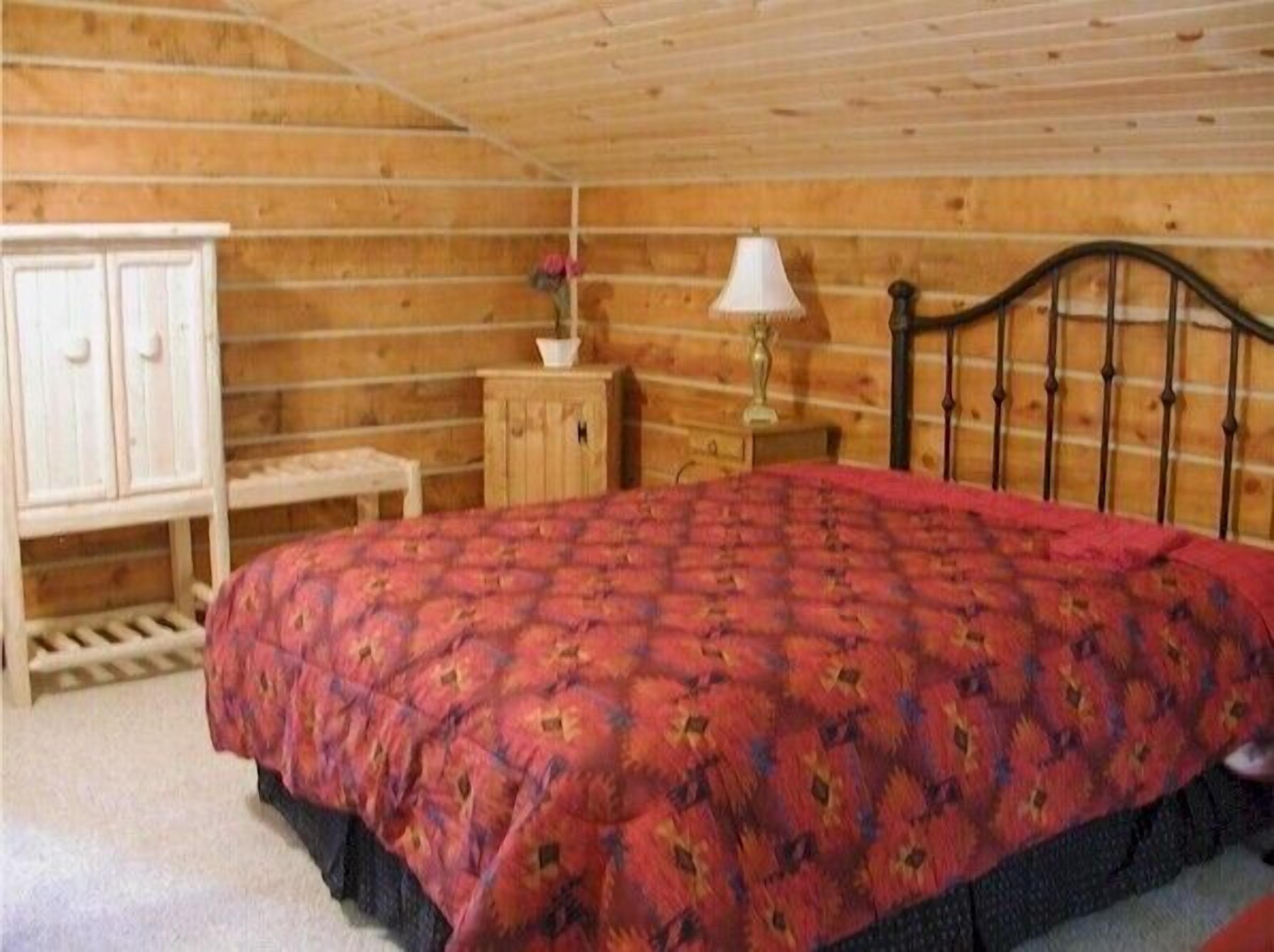 Une chambre confortable avec un plafond en bois, un lit recouvert d’une courtepointe à motifs rouges, une armoire blanche et une petite table de chevet avec une lampe.