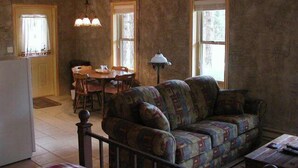 A rustic living room with a patterned sofa, a dining table with chairs, and a kitchen area with a refrigerator.