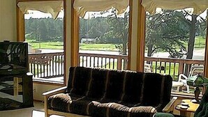 A room with a wooden bench, a small table with a lamp, and a view of a green landscape through large windows. - Thunderheart (Custer)