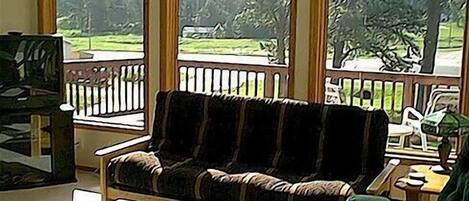 A room with a wooden bench, a small table with a lamp, and a view of a green landscape through large windows.