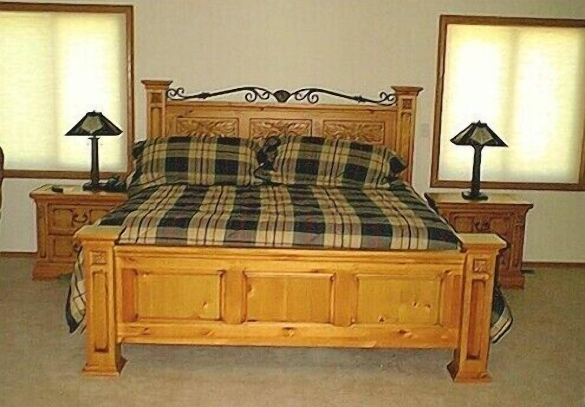 Una cama de madera con cabecera, flanqueada por dos mesitas de noche con lámparas, en una habitación con paredes lisas y ventanas.