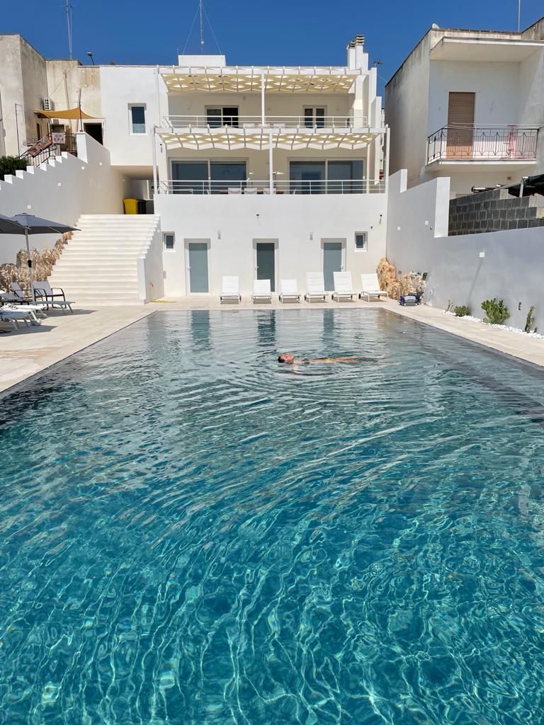 Ein Schwimmbecken mit klarem Wasser, umgeben von weißen Gebäuden mit Balkonen und einer Treppe, die zu einer erhöhten Ebene führt.