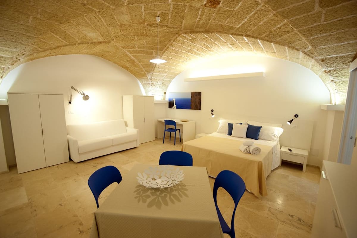 A cozy, well-lit room with a stone ceiling, a white sofa, a bed with blue pillows, a small table with a decorative centerpiece, and a chair.