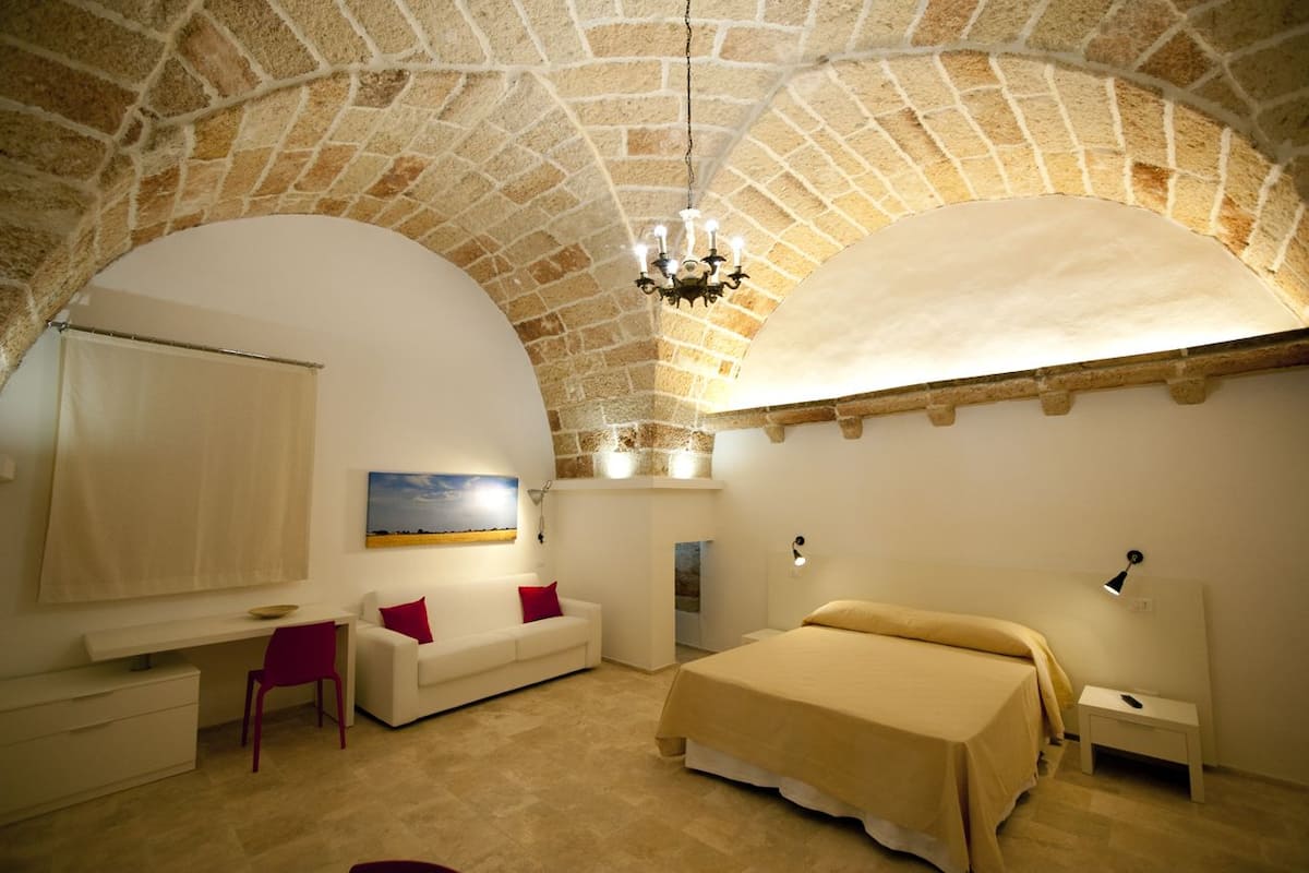 A room with a stone ceiling, a bed, a desk, a chair, and a painting on the wall.