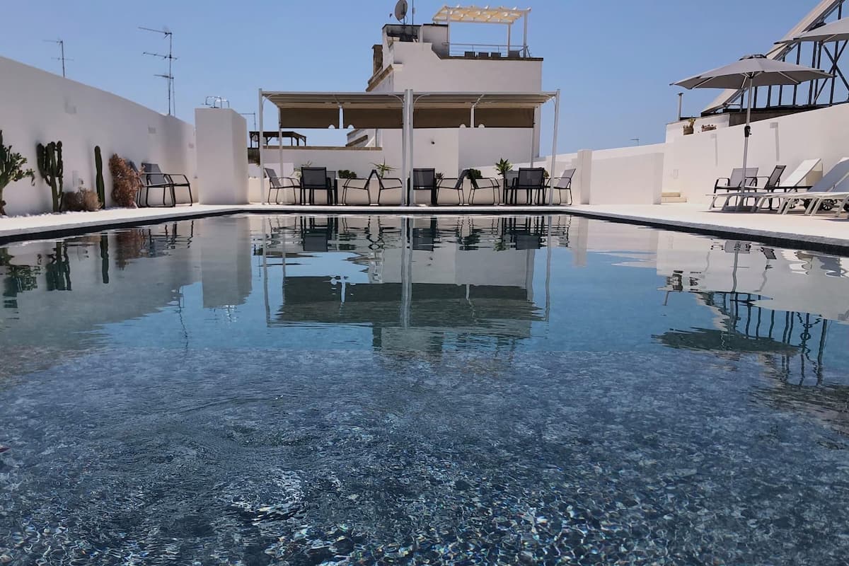 A clear swimming pool with lounge chairs and umbrellas, surrounded by white walls and a building with a flat roof.