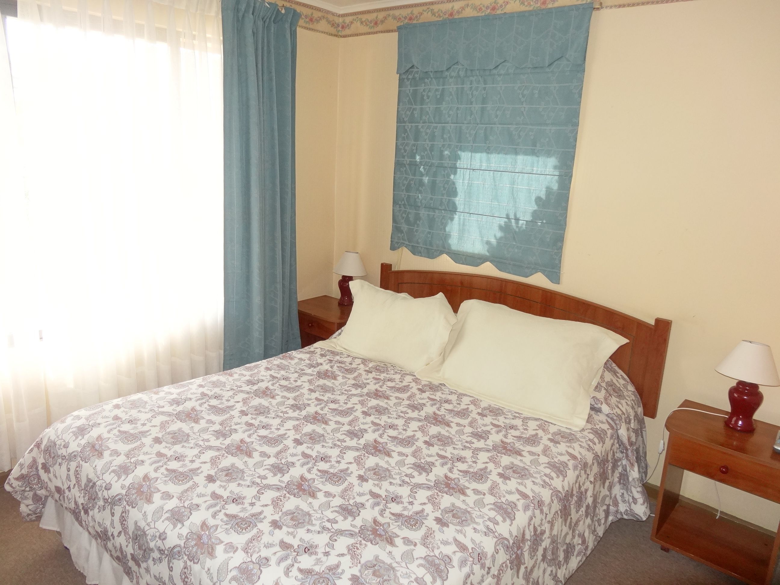 Un dormitorio con cama, dos mesitas de noche, una mesita auxiliar, una ventana con cortinas y un adorno en la pared.