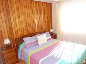 Een slaapkamer met een houten hoofdbord, een bed met beddengoed met een patroon, twee nachtlampjes en een nachtkastje met een afstandsbediening.