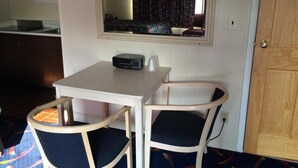 Ein kleines Zimmer mit einem weißen Tisch, zwei Stühlen, einem Spiegel über dem Tisch und einer Tür rechts.