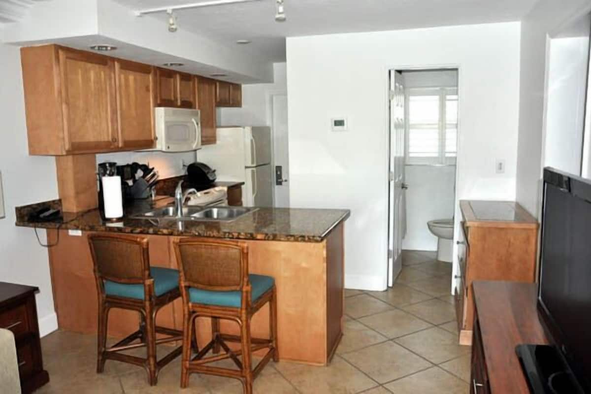 A kitchen with wooden cabinets, a microwave, a refrigerator, and a sink. There are two chairs with blue cushions.