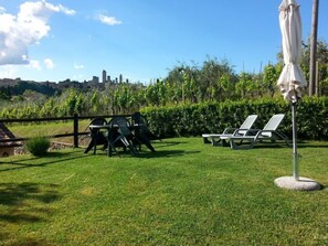A grassy outdoor area with a table and chairs, a white umbrella, and a view of a cityscape in the background.