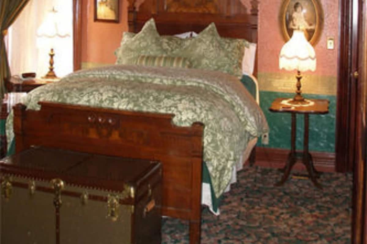 A room with a wooden bed, a green upholstered chair, a small table, and a wall-mounted lamp.