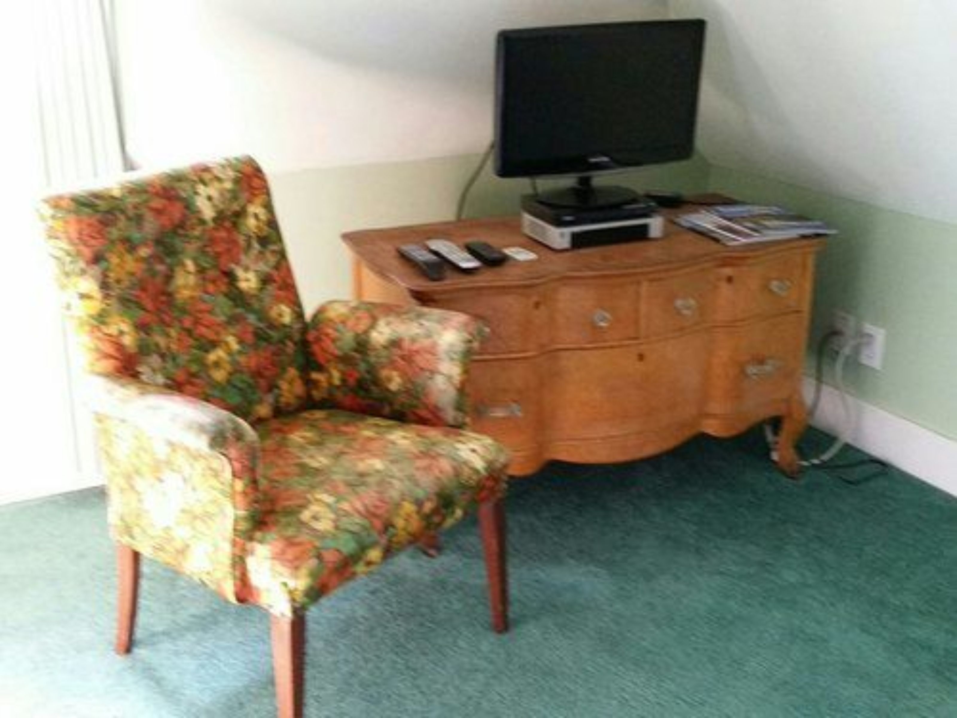 A room with a floral-patterned armchair, a wooden dresser with a flat-screen TV, and a green carpet.