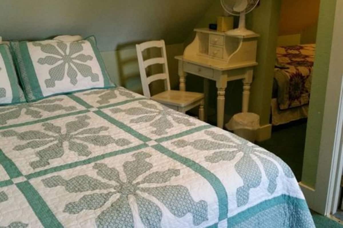 A room with a bed, a chair, a small table with a fan, and a door leading to another room.