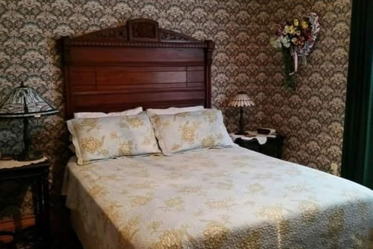 A bedroom with a wooden headboard, floral patterned bedding, a nightstand with a lamp, and a wall-mounted flower arrangement.