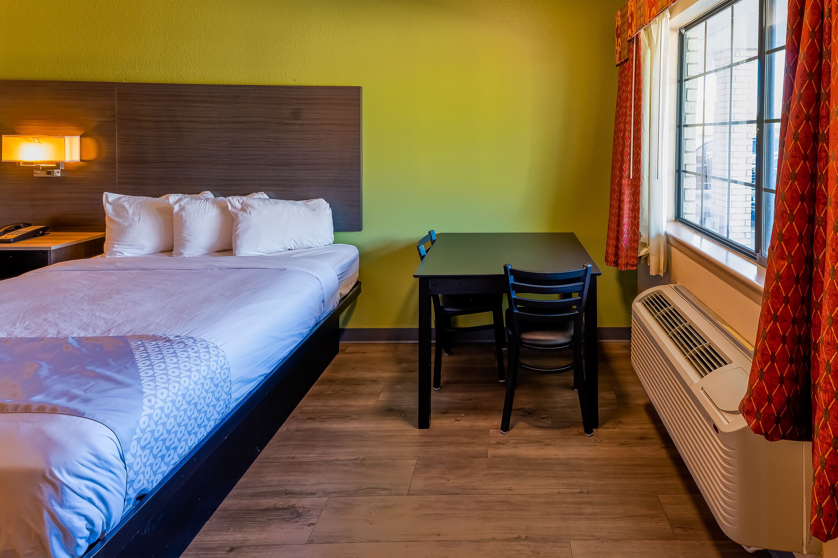 A hotel room with a bed, a desk, two chairs, a heater, and a window with curtains.