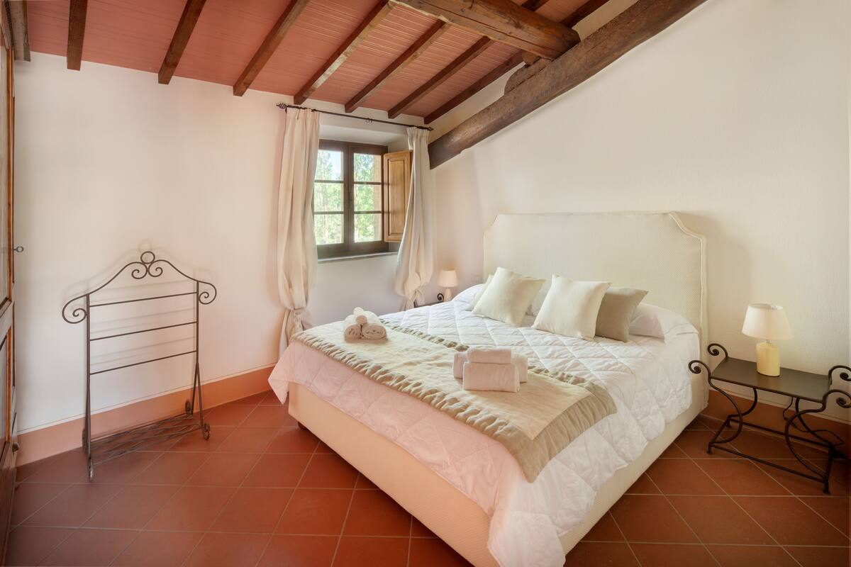 A bedroom with a bed, a nightstand, a window with curtains, a metal rack, and a wooden ceiling.