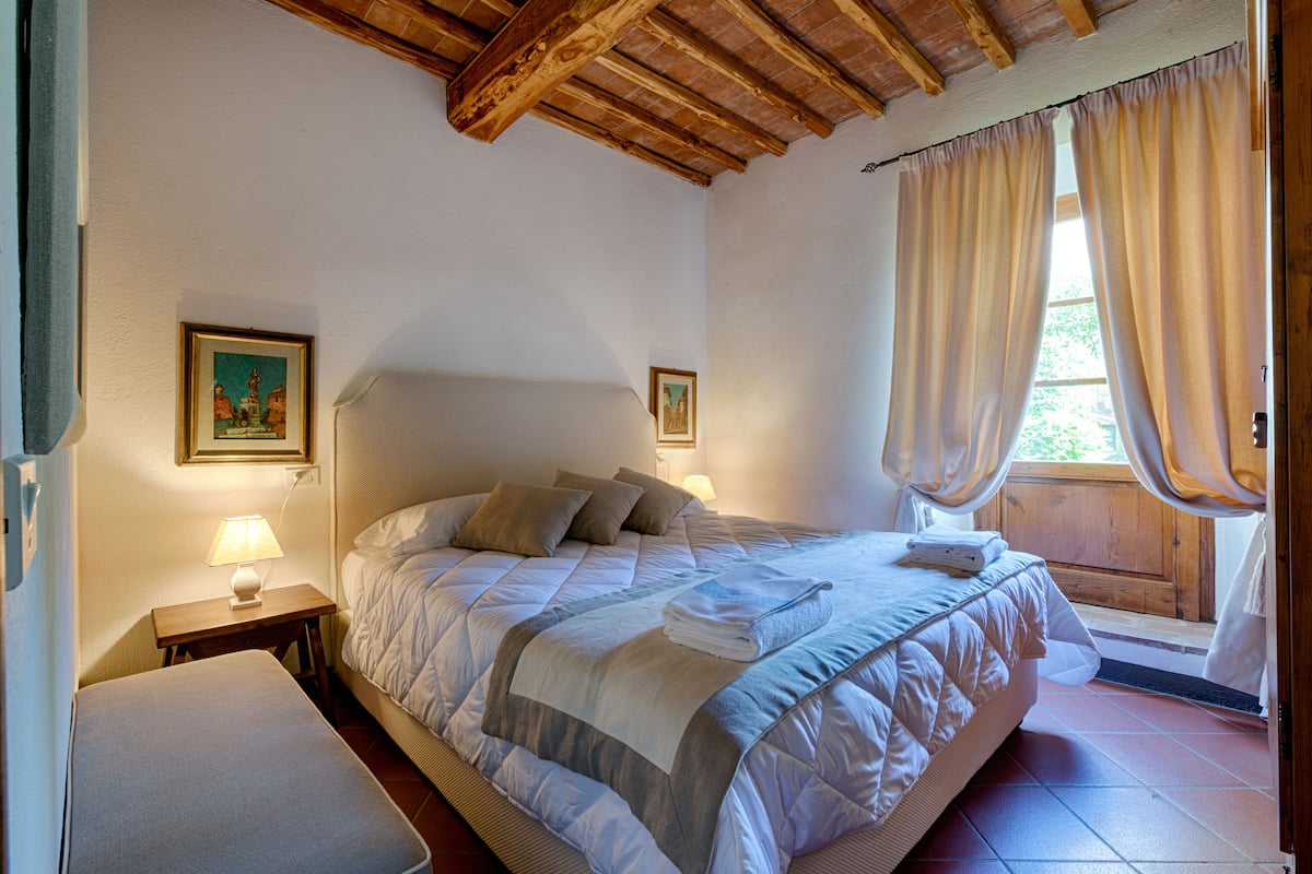 A bedroom with a large bed, two bedside tables with lamps, a window with curtains, and wooden beams on the ceiling.