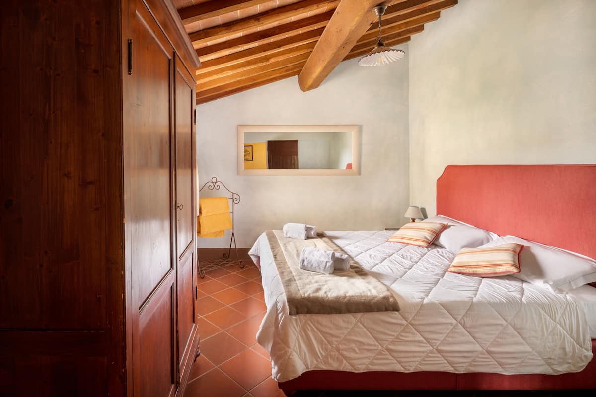 A bedroom with a wooden wardrobe, a bed with white bedding and striped pillows, a red headboard, and a small bedside table with a lamp.