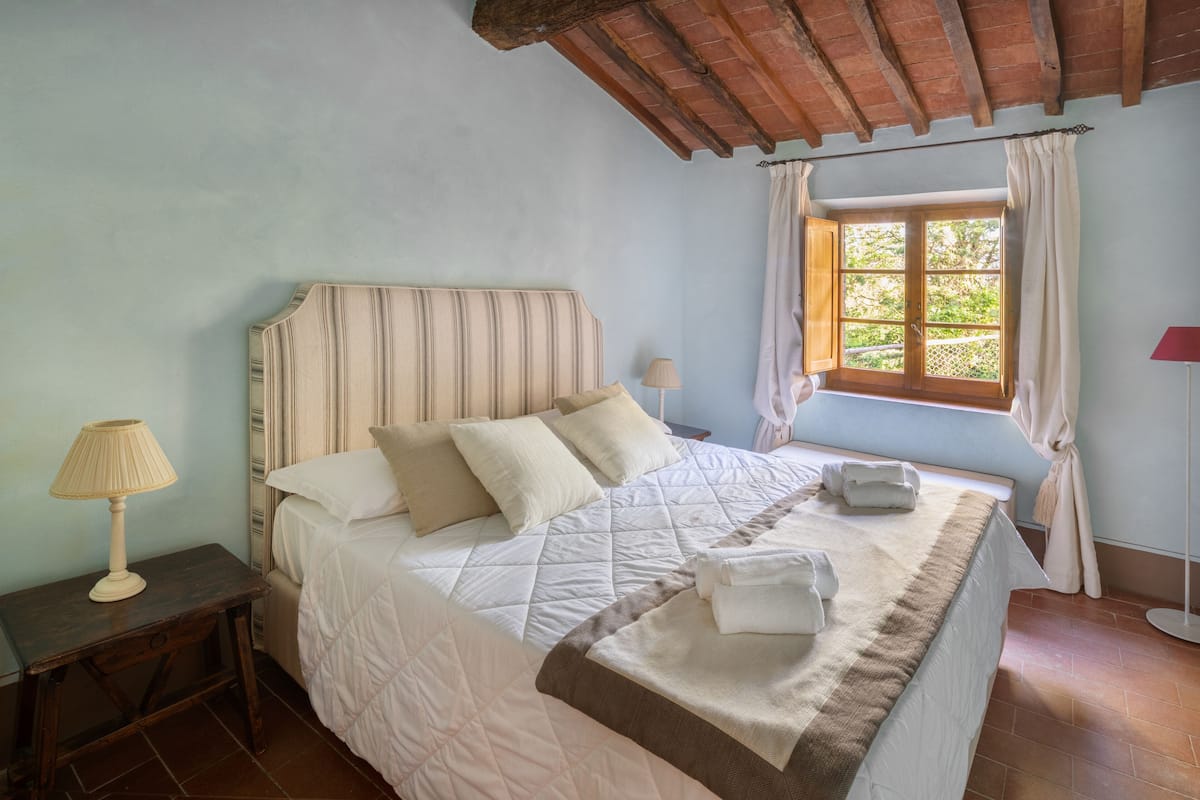 A bedroom with a bed, a nightstand, a lamp, a window with curtains, and a view of greenery outside.