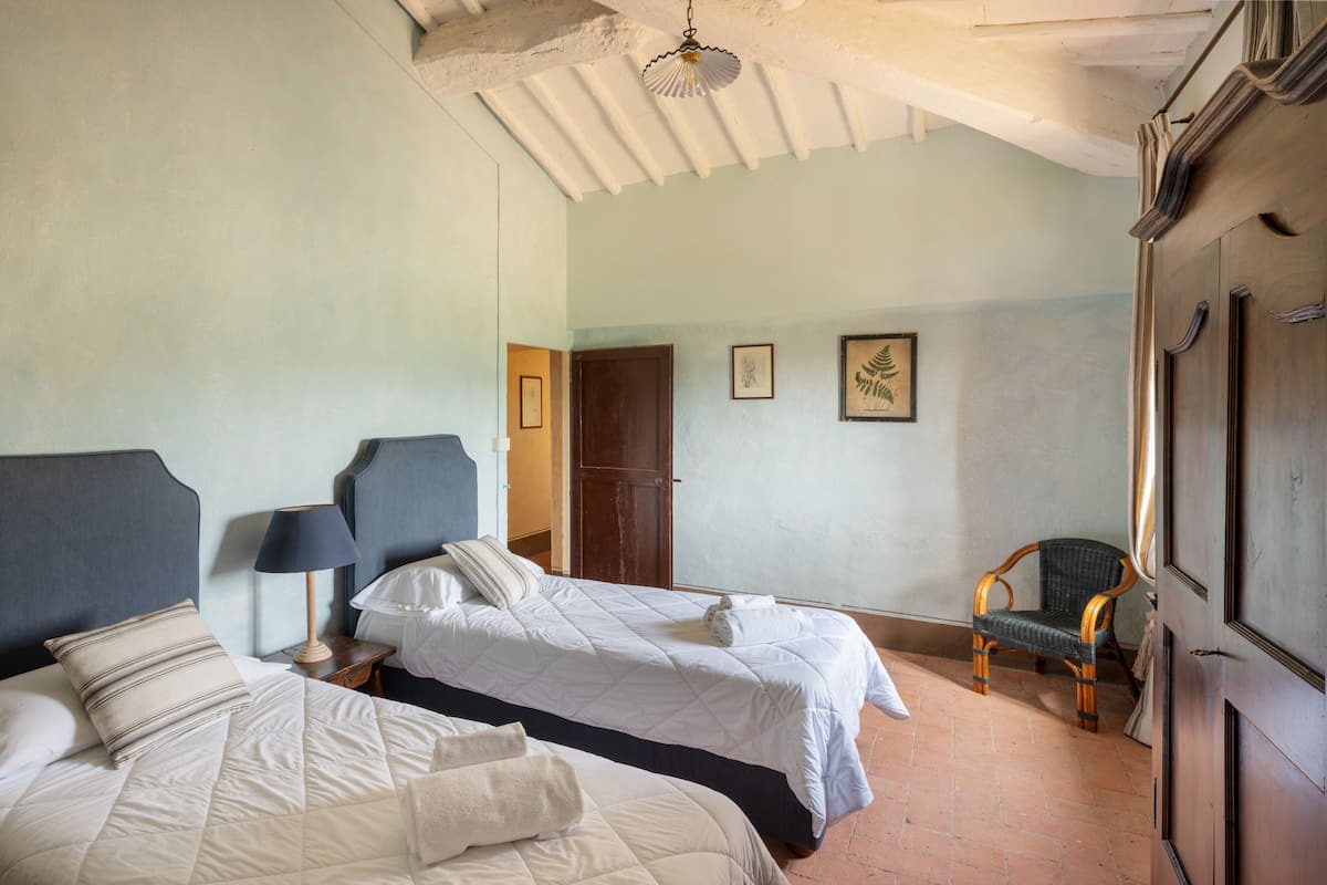 A room with two beds, a chair, a door, and a ceiling with exposed beams.