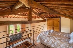 A bedroom with a wooden ceiling, a bed with white bedding, a wooden door, and a window with a view of greenery.