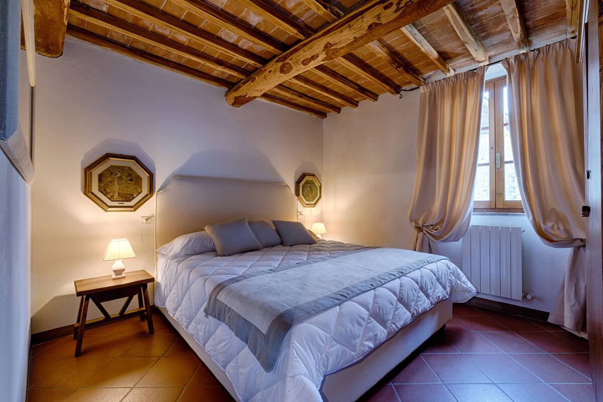 A bedroom with a wooden ceiling, a bed with a white duvet, two bedside tables with lamps, and a window with curtains.