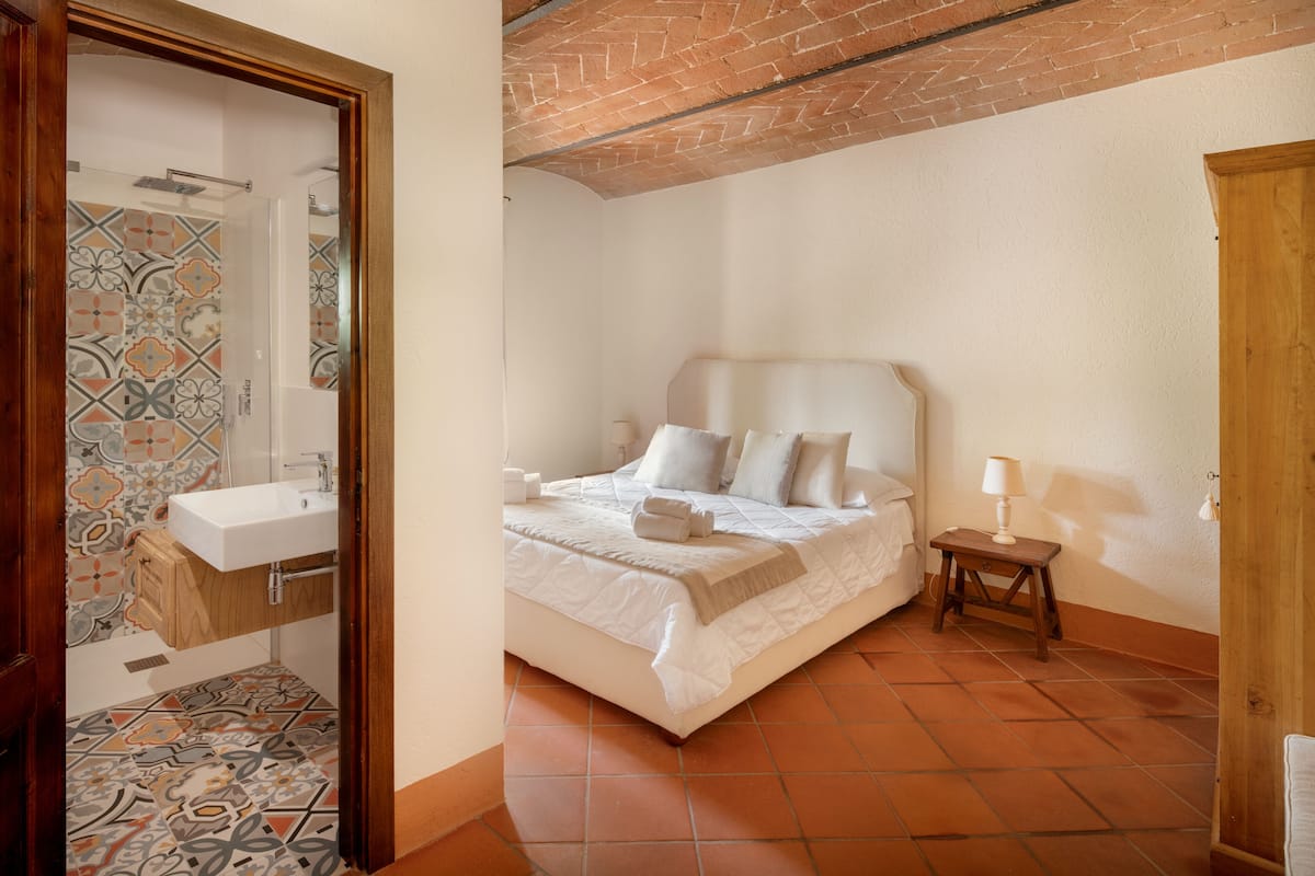 A bedroom with a bed, bedside table, and a bathroom with a sink and shower.