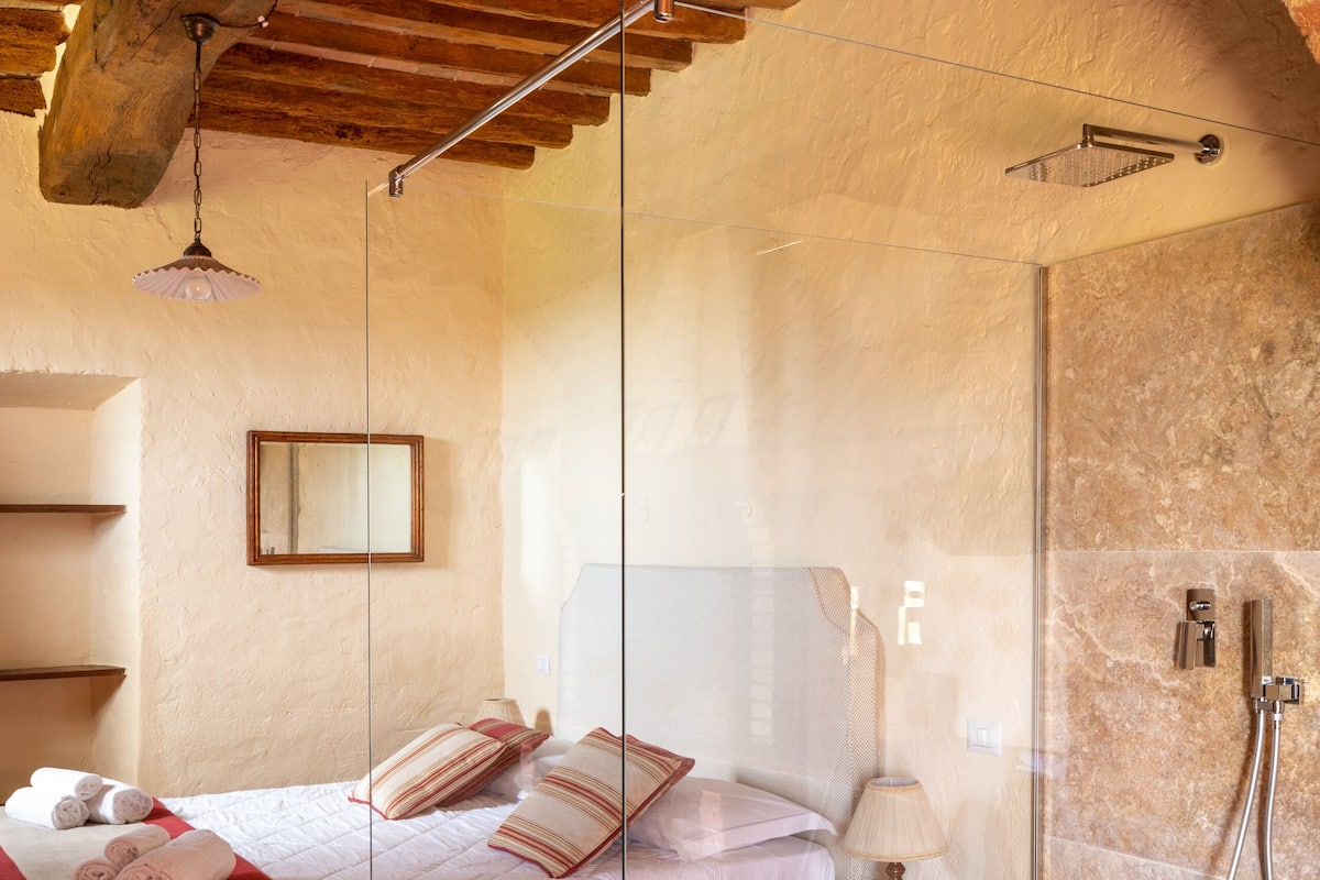 A bedroom with a bed, a bedside table, a lamp, and a shower area with a glass door.