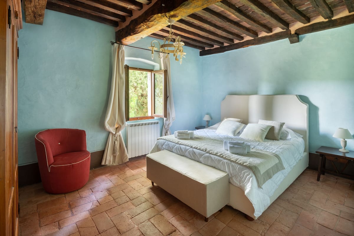 A bedroom with a large bed, a red chair, a window with curtains, and a wooden ceiling.
