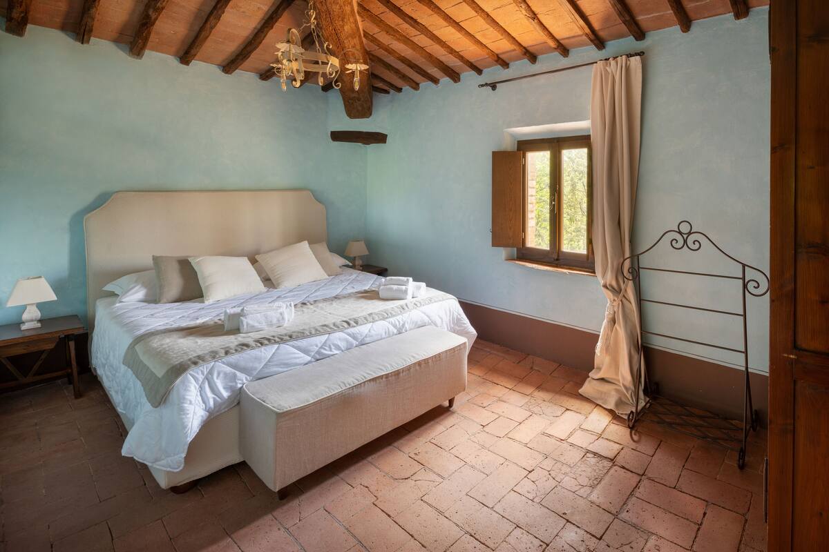 A bedroom with a large bed, a wooden ceiling, a window with curtains, and a nightstand with a lamp.