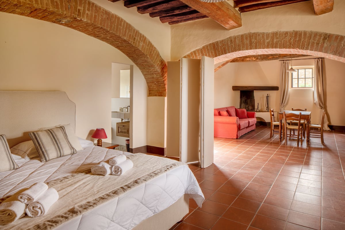 A hotel room with a large bed, a red sofa, a dining table with chairs, and a fireplace.