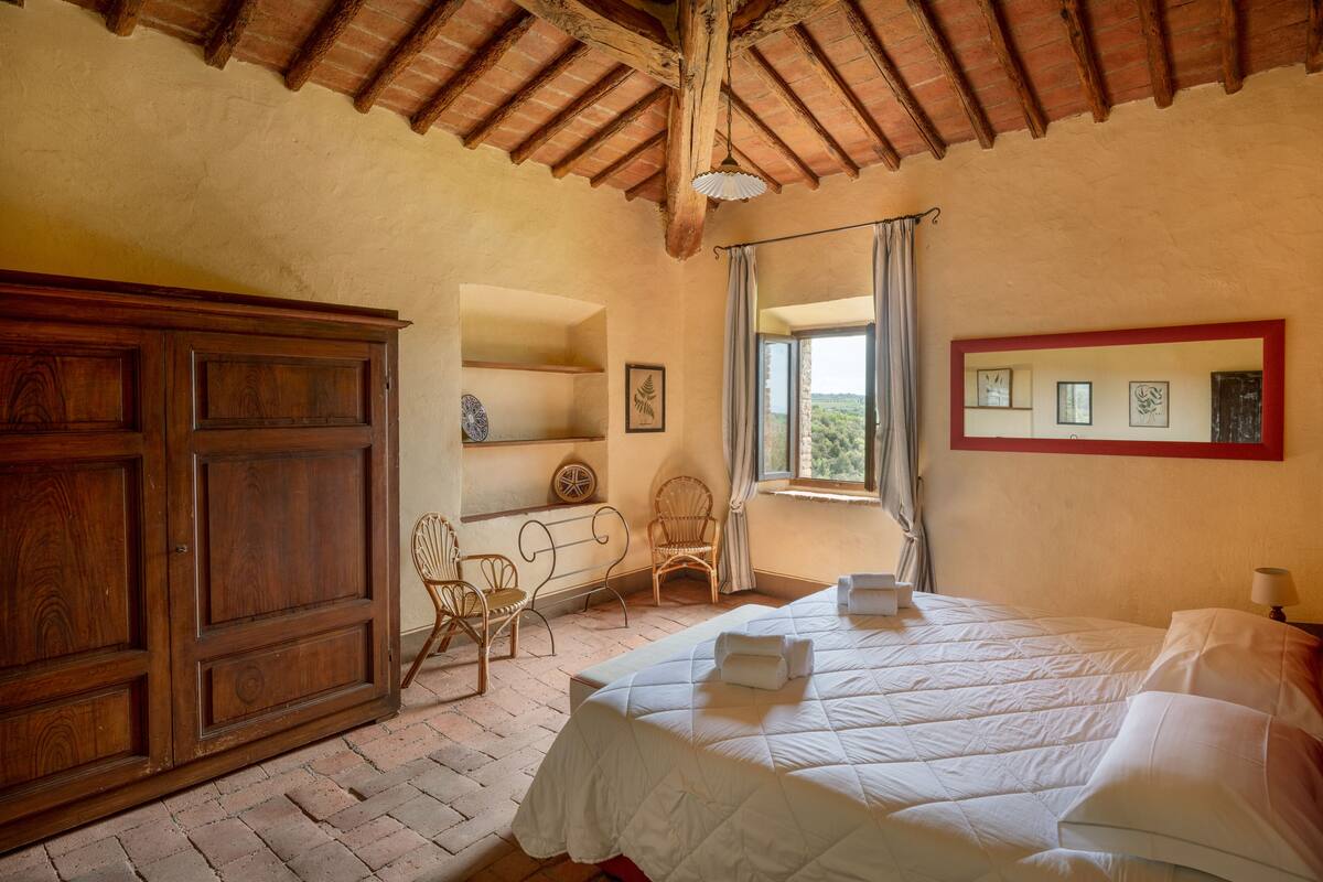 A bedroom with a wooden wardrobe, a bed with white linens, a wicker chair, and a window with curtains.