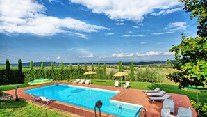 Piscina com espreguiçadeiras e guarda-sóis, cercada por um jardim bem cuidado e vista para colinas ao longe.