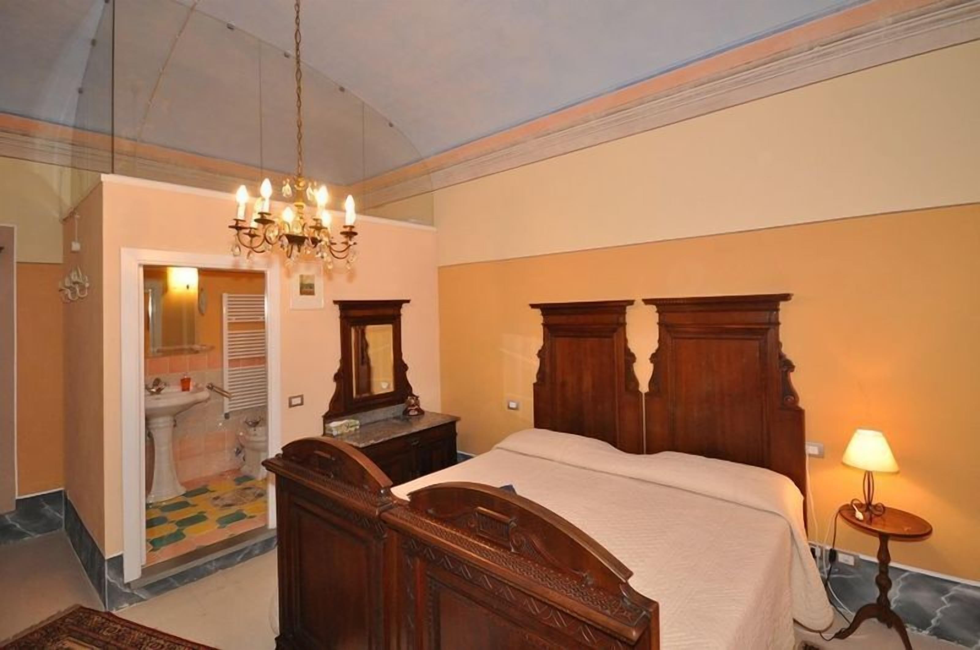 A bedroom with a wooden bed, a chandelier, a bedside lamp, and a bathroom visible through an open door.