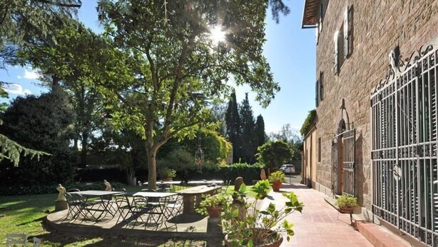 Un patio con mobiliario exterior, un árbol grande y un edificio de piedra con una verja de metal.
