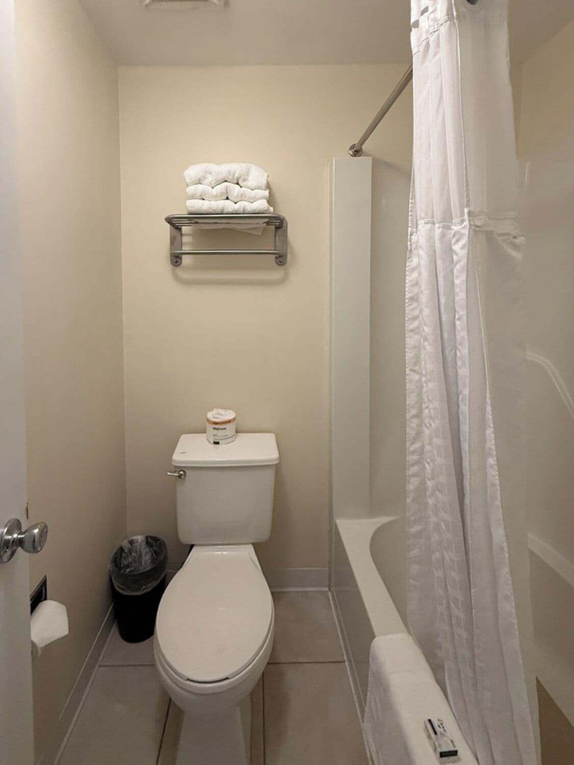 Een badkamer met een toilet, een plank met handdoeken, een prullenbak en een douchegordijn.