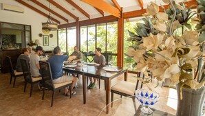 Uma área de refeições com mesa e cadeiras de madeira, janelas amplas e um arranjo de flores pendente.