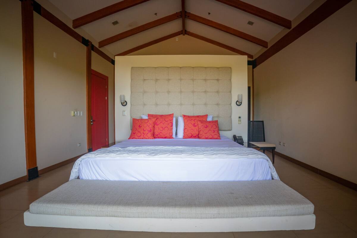 A large bed with a headboard, red pillows, and a bedside table with a lamp.
