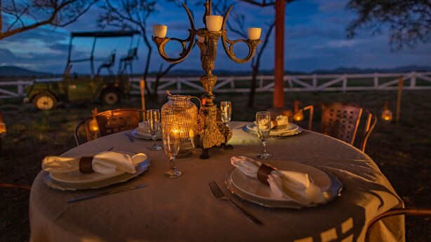 Mesa de jantar ao ar livre posta para refeição, com candelabro como peça central, rodeada por cadeiras e um cercado branco ao fundo.