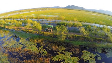 Vista aérea de un paisaje con un vehículo verde estacionado cerca de una masa de agua rodeada de vegetación y una cadena montañosa distante.