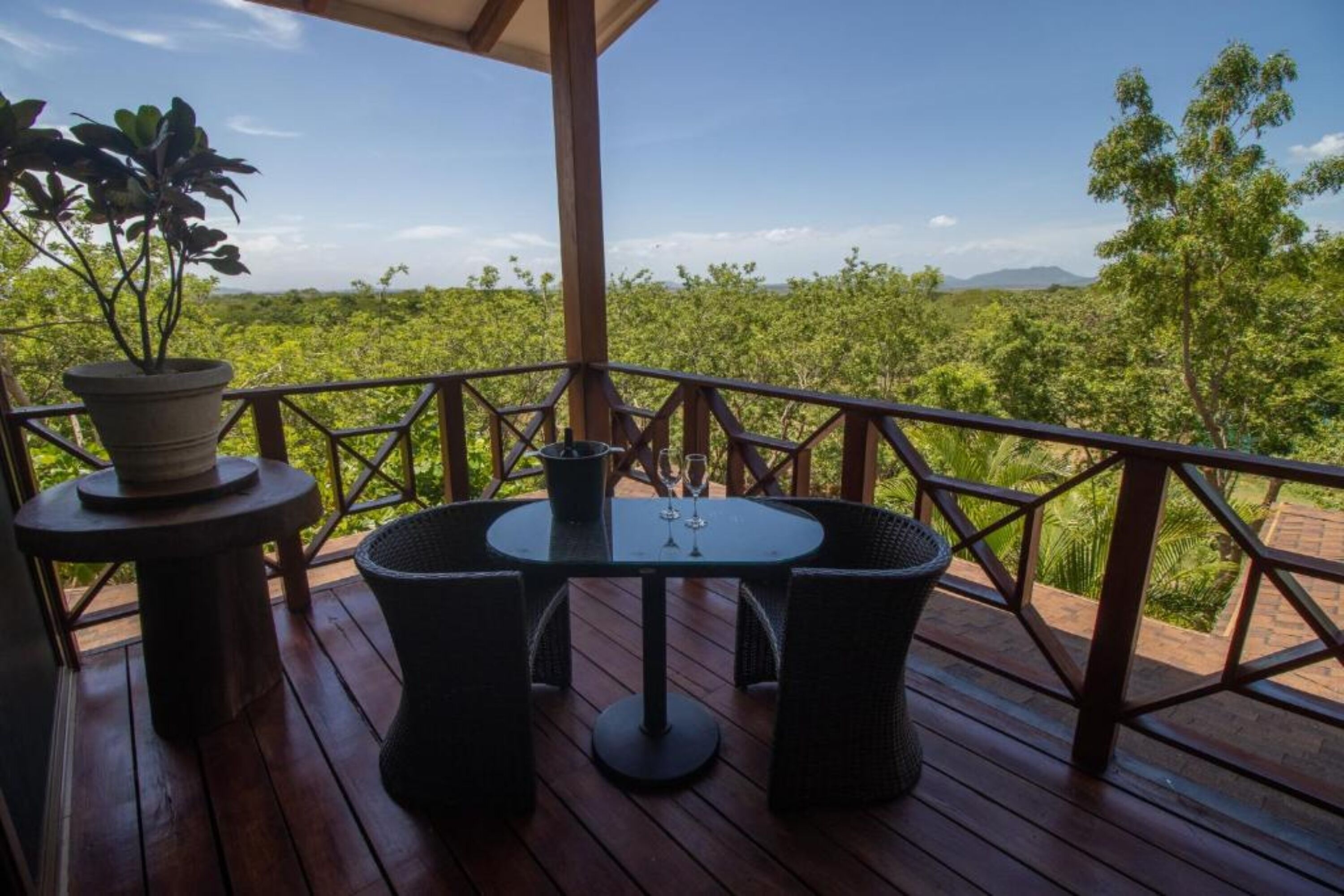 Una terraza de madera con una mesa redonda y sillas, una planta en maceta y vistas a un paisaje verde y exuberante.