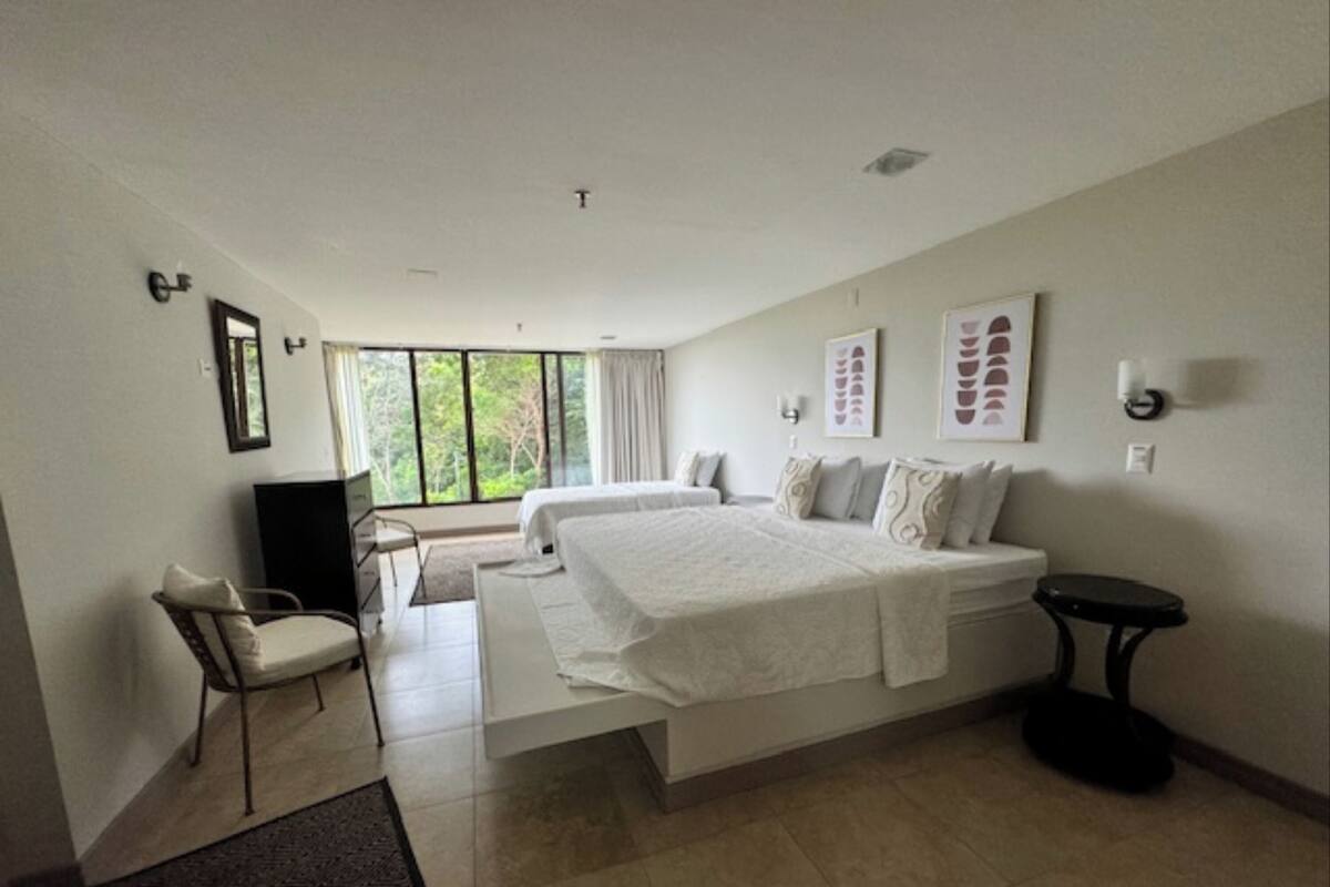 A modern hotel room with a large bed, a desk, and a chair. There is a window with a view of trees.